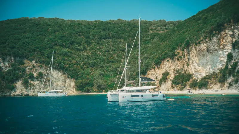 A cruising catamaran anchored in a turquoise cove below green cliffs.